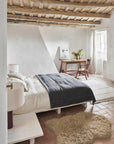 A minimalist bedroom featuring rustic wooden ceiling beams, a cozy bed with a dark gray blanket, a Lorena Canals Woolable Sheepskin Rug on terracotta tile flooring, and a wooden desk with a chair near a window. The space is decorated with light, neutral tones.