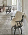 A minimalist room with two wooden chairs, one draped with a white fluffy throw, positioned on a gray Woolable Sheepskin Rug by Lorena Canals. A wooden desk and chair are situated near a window, with light streaming in. A simple vase with flowers and framed artwork are visible on the desk and wall, emphasizing an eco-friendly aesthetic.