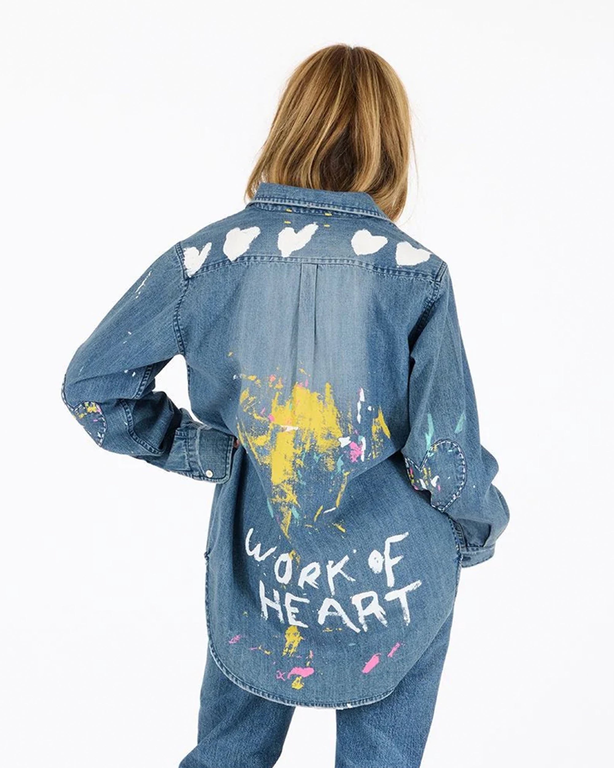 A person with long hair, seen from behind, wears the Kerri Rosenthal Mia Studio Shirt—a painted denim shirt with white hearts on the shoulders, colorful paint splatters, and "WORK OF HEART" hand-painted on the back.