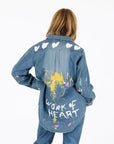 A person with long hair, seen from behind, wears the Kerri Rosenthal Mia Studio Shirt—a painted denim shirt with white hearts on the shoulders, colorful paint splatters, and "WORK OF HEART" hand-painted on the back.