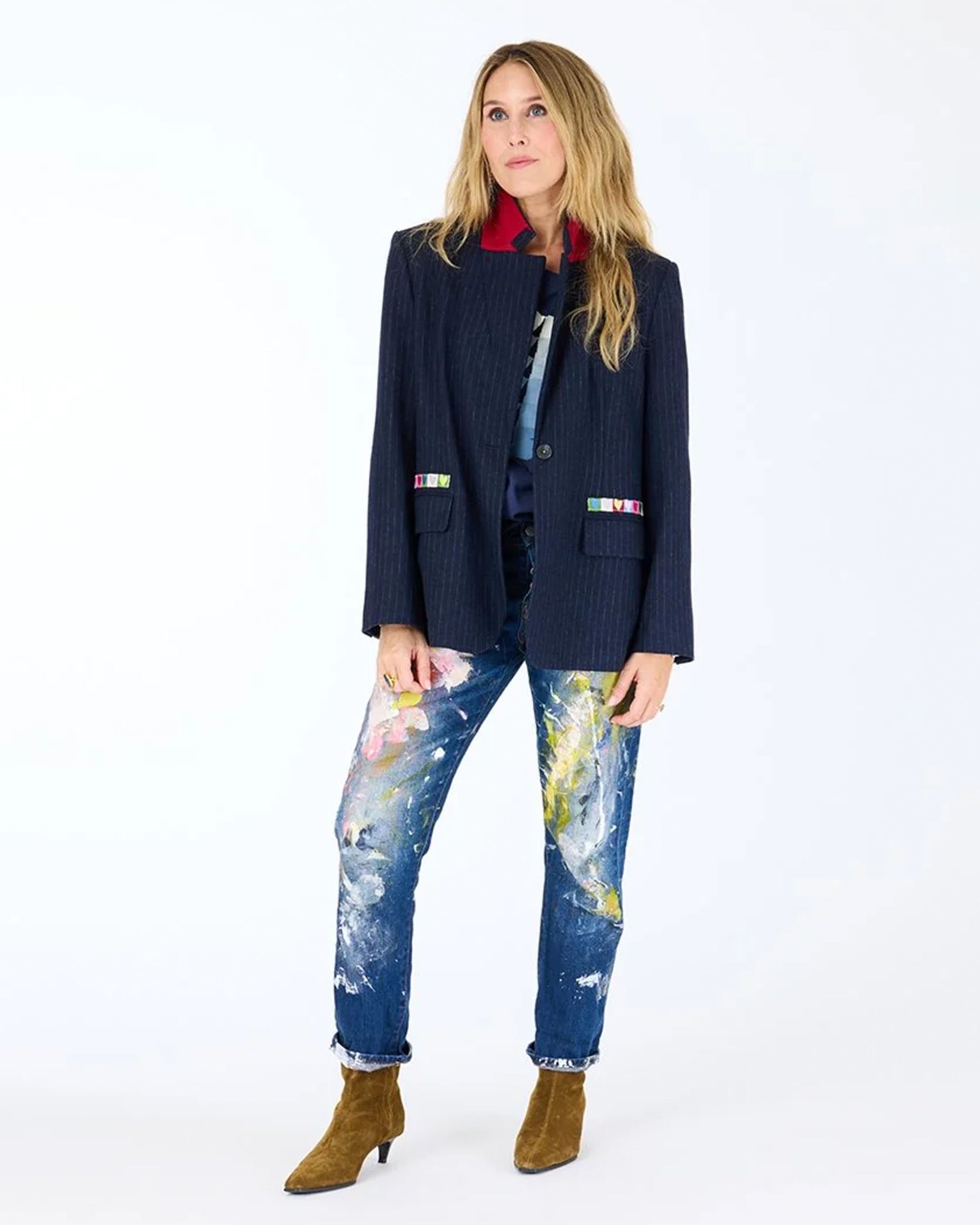 A woman with long blonde hair stands against a white background, wearing the Kerri Rosenthal Workday Love Brigade Blazer in navy pinstripe, a red shirt, paint-splattered jeans, and brown ankle boots. She gazes to the side with a neutral expression.