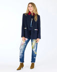 A woman with long blonde hair stands against a white background, wearing the Kerri Rosenthal Workday Love Brigade Blazer in navy pinstripe, a red shirt, paint-splattered jeans, and brown ankle boots. She gazes to the side with a neutral expression.