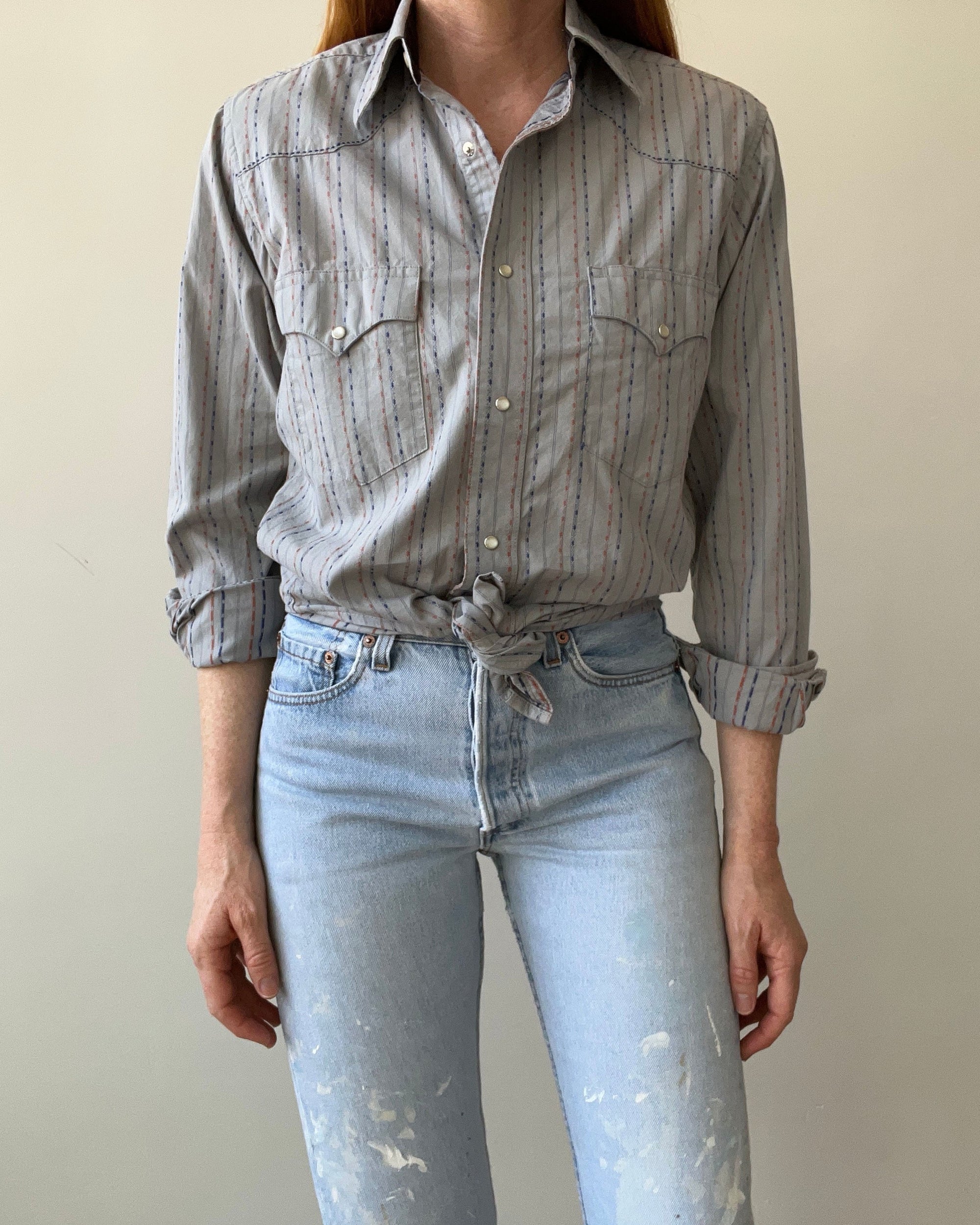 A person wears the 80s Wrangler Button Up from Red Vintage Co., sleeves rolled and tied at the waist, paired with high-waisted light blue jeans with white paint splatters.