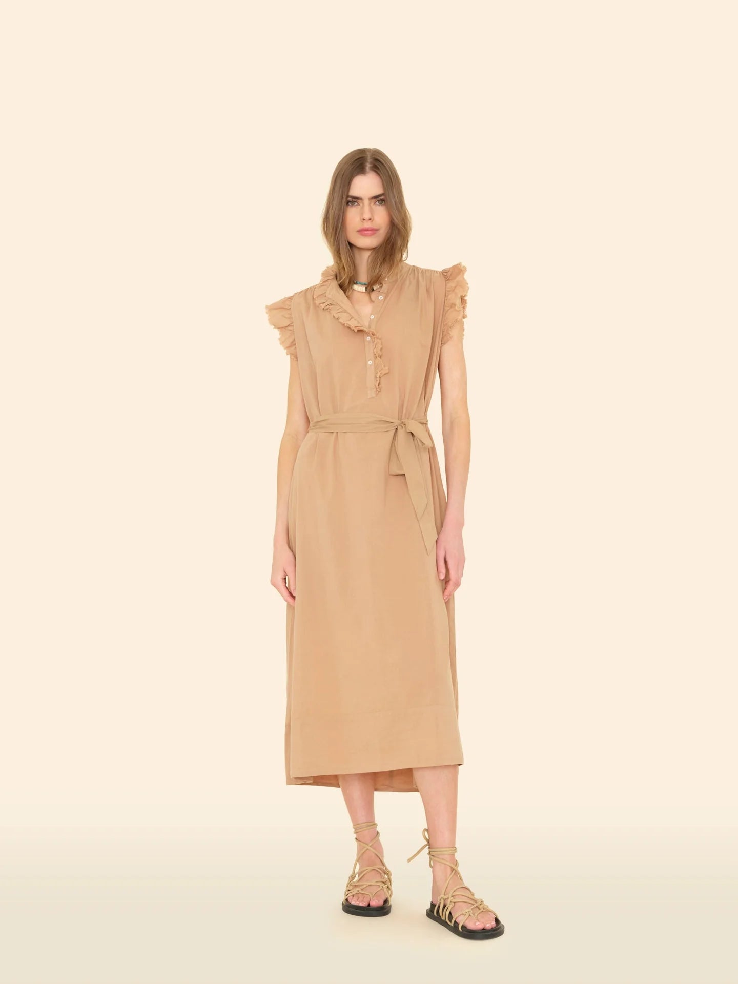 A person stands against a plain background in the Xirena April Dress, a light brown cotton poplin midi with shoulder ruffles and a matching tie belt. They have long hair and wear strappy flat sandals.