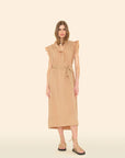 A person stands against a plain background in the Xirena April Dress, a light brown cotton poplin midi with shoulder ruffles and a matching tie belt. They have long hair and wear strappy flat sandals.