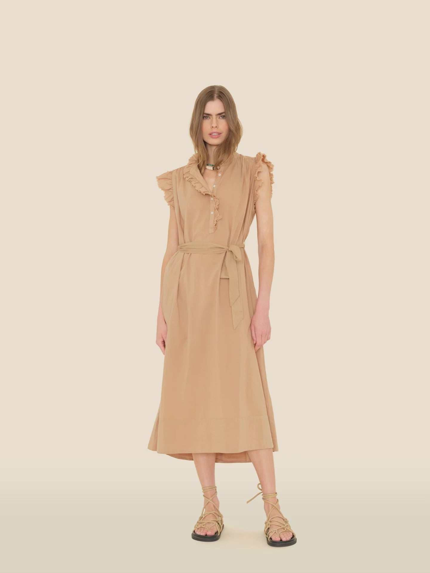 A person wears the Xirena April Dress, a tan sleeveless midi made of cotton poplin, featuring ruffle details and a waist tie. They're paired with strappy sandals against a light beige backdrop.