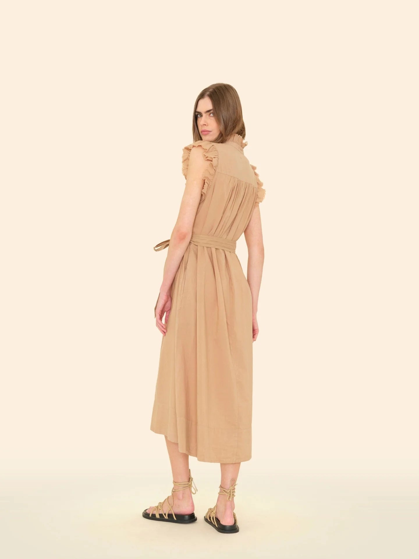 A person with long hair stands in profile against a beige background, wearing the April Dress by Xirena. This sleeveless, knee-length beige midi dress is made of cotton poplin with ruffle details and a tied waist. They are also wearing strappy beige sandals.