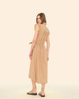 A person with long hair stands in profile against a beige background, wearing the April Dress by Xirena. This sleeveless, knee-length beige midi dress is made of cotton poplin with ruffle details and a tied waist. They are also wearing strappy beige sandals.