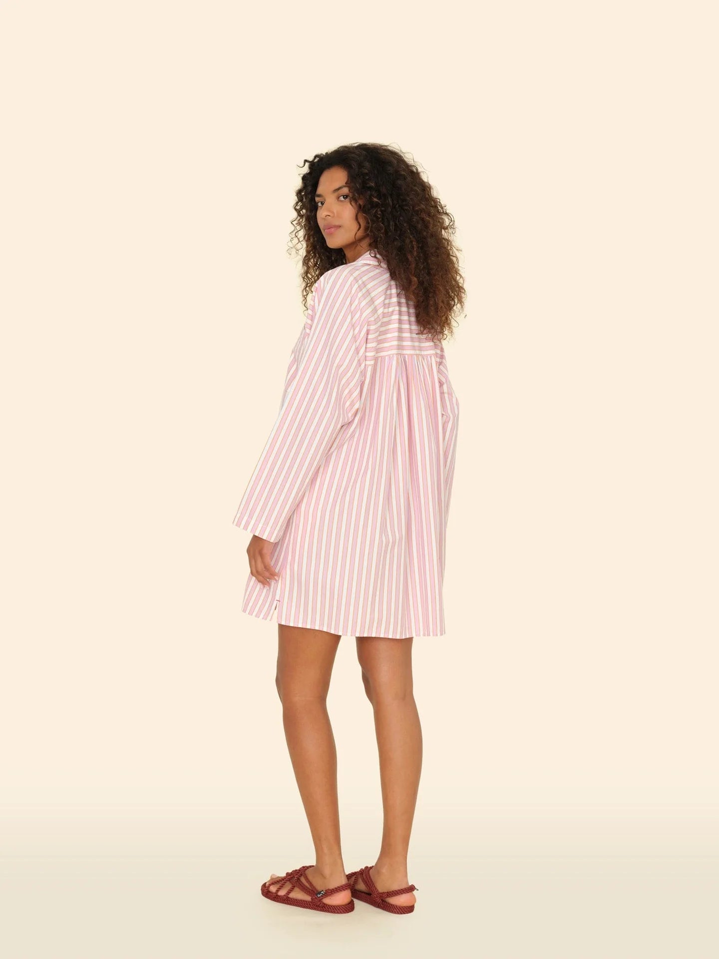 With their charming curly hair, a person dons Xirena's Flora Dress, pink with white stripes and 100% cotton, paired with brown sandals. They stand against a beige backdrop, glancing over their shoulder.