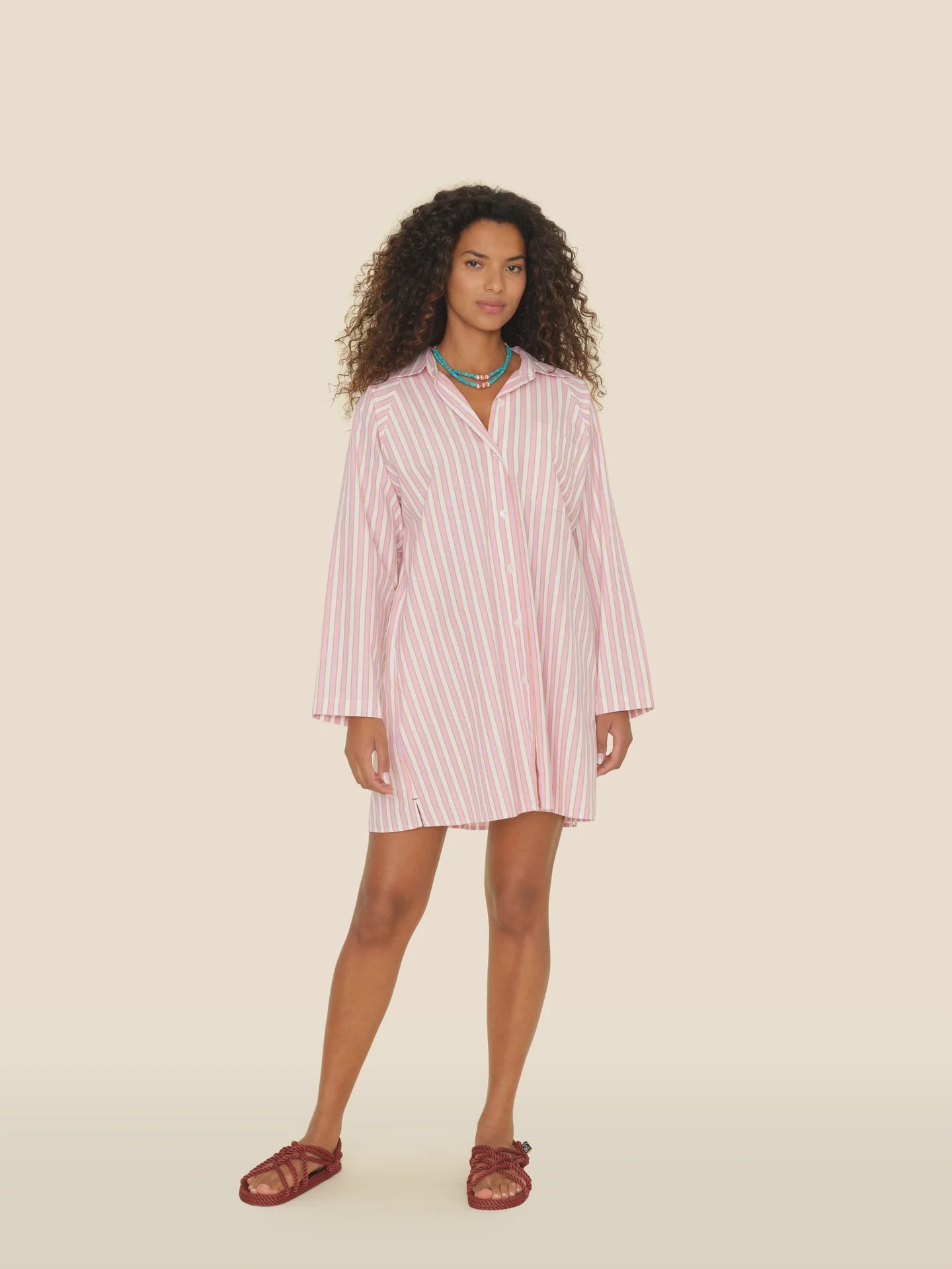 A woman in a Flora Dress by Xirena stands against a beige background. Her 100% cotton outfit perfectly complements her curly hair, green necklace, and red sandals.