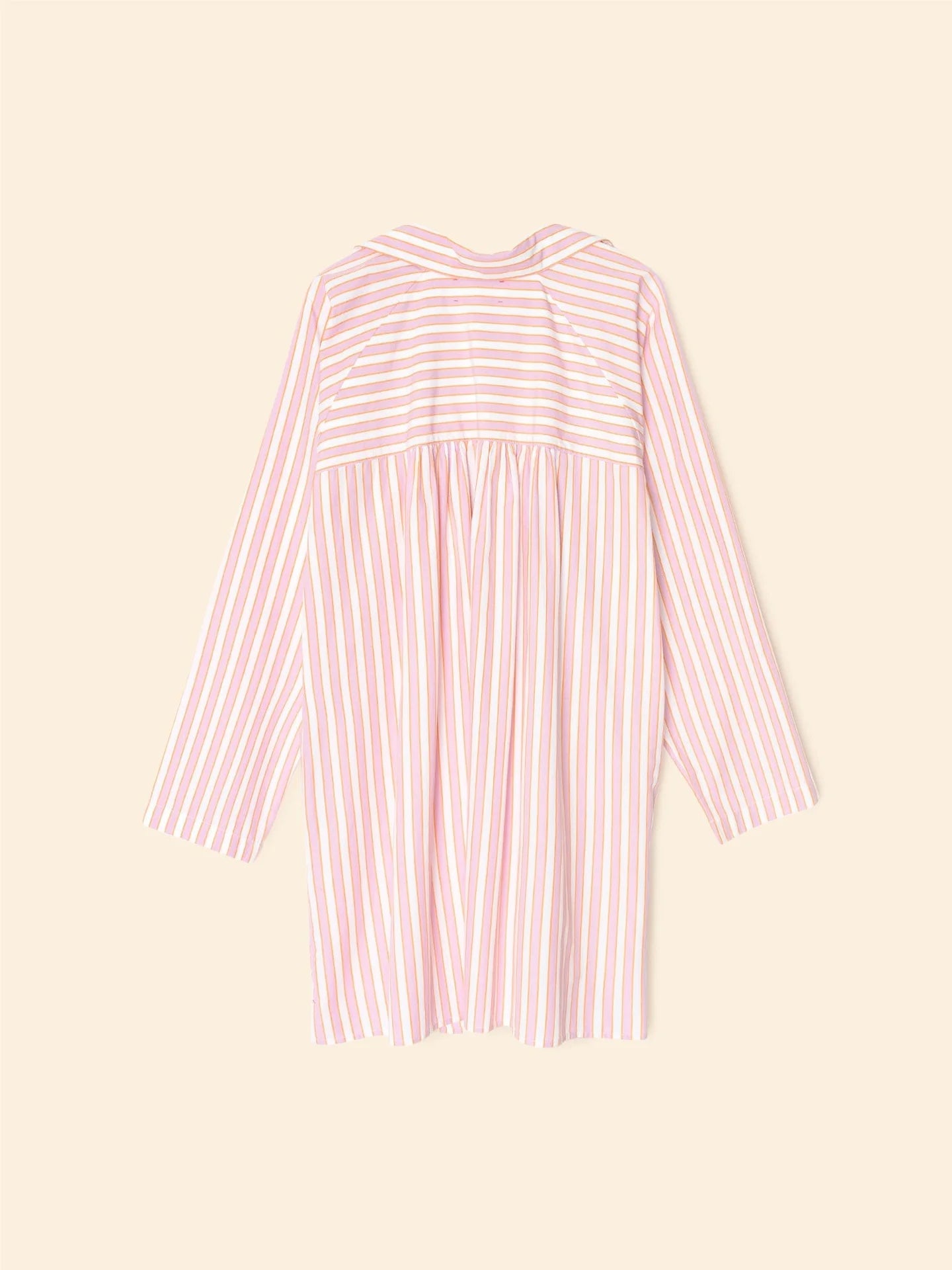 Introducing the Flora Dress by Xirena: a mini shirtdress featuring pink and white vertical stripes. Crafted from 100% cotton, it includes long sleeves, a yoke with horizontal stripes, and a gathered section below against a plain beige background.