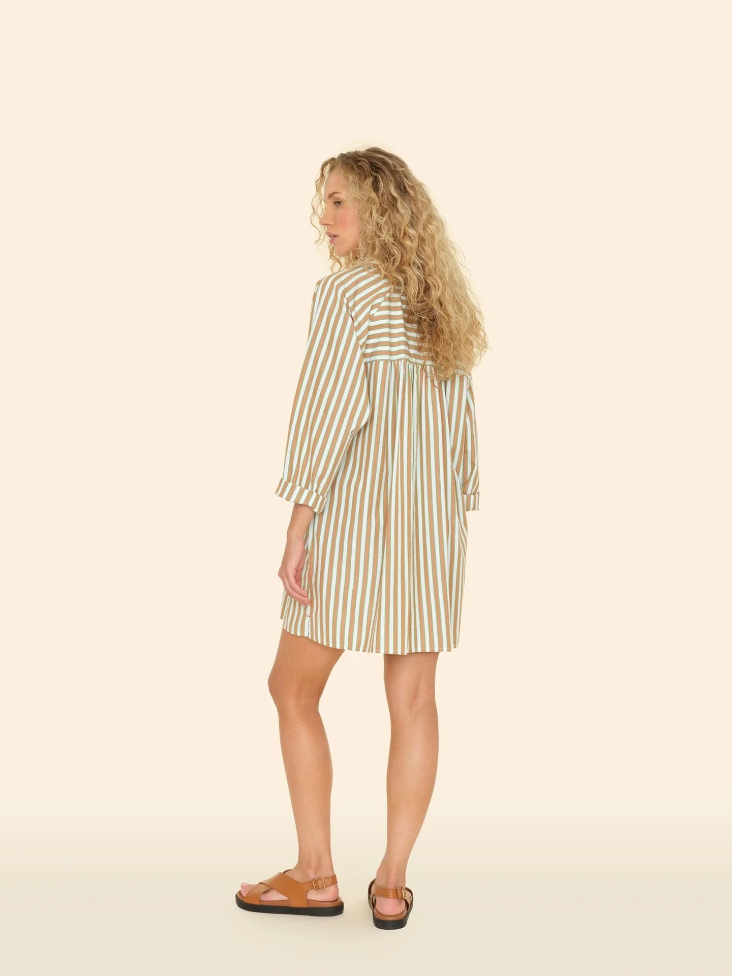 A person with curly hair wears a Flora Dress by Xirena and sandals, facing away. The 100% cotton dress showcases vertical white and brown stripes on a light beige background.