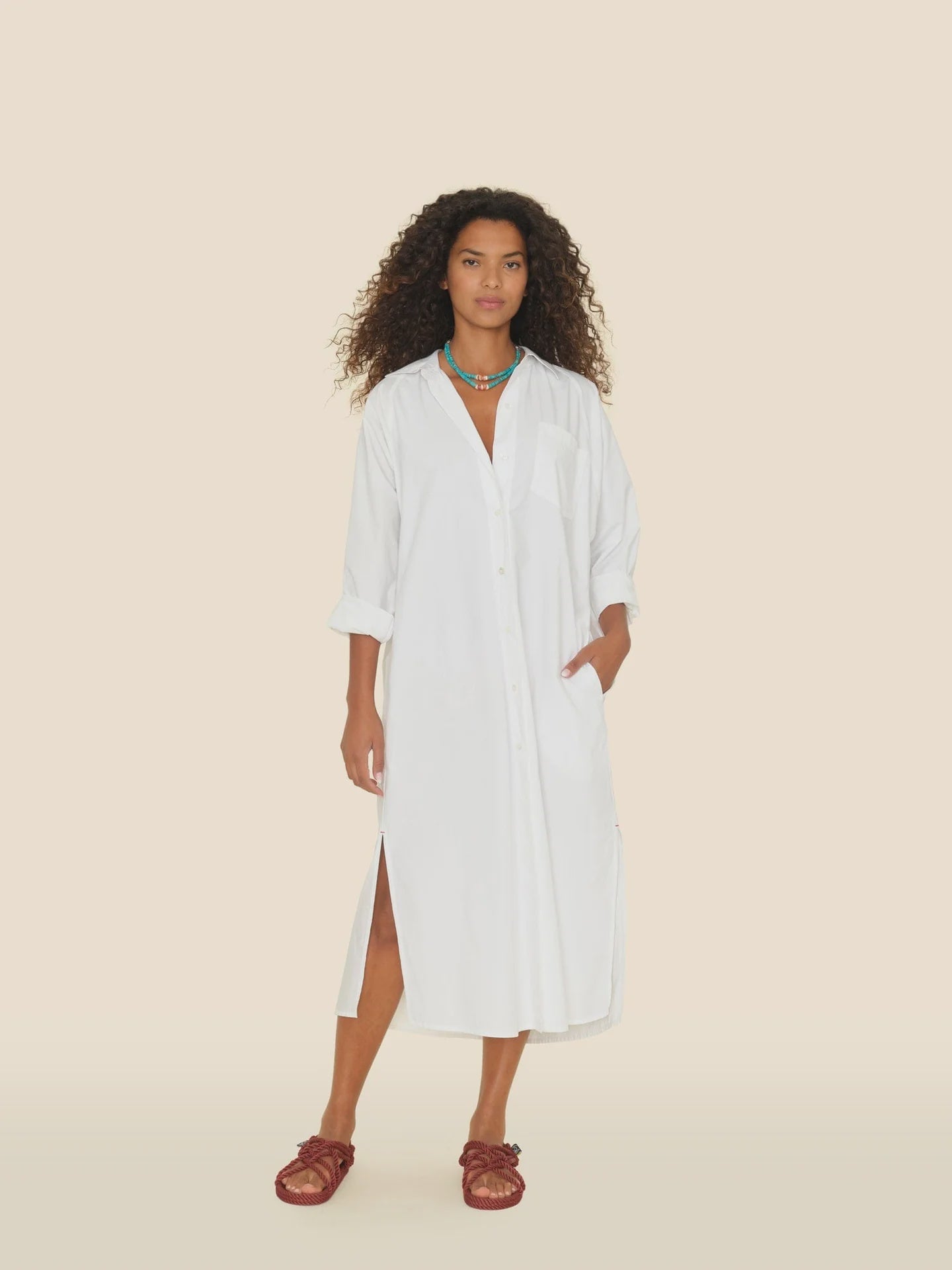 A woman with curly hair stands against a beige background, wearing a Xirena Marfa Dress made from crisp paper poplin. The long white button-up dress features rolled sleeves and is accessorized with one hand in a pocket, red sandals, and colorful beaded necklaces.