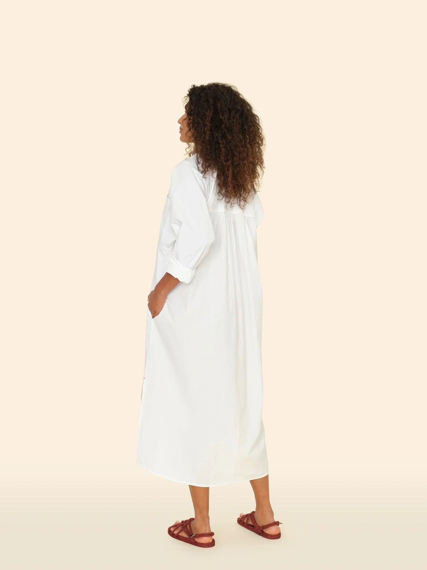 A woman with curly hair stands facing away in the Marfa Dress by Xirena, a relaxed shirtdress made from crisp paper poplin, paired with brown sandals. She gazes slightly to the side against a plain beige background.