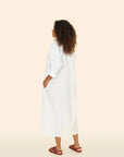 A woman with curly hair stands facing away in the Marfa Dress by Xirena, a relaxed shirtdress made from crisp paper poplin, paired with brown sandals. She gazes slightly to the side against a plain beige background.