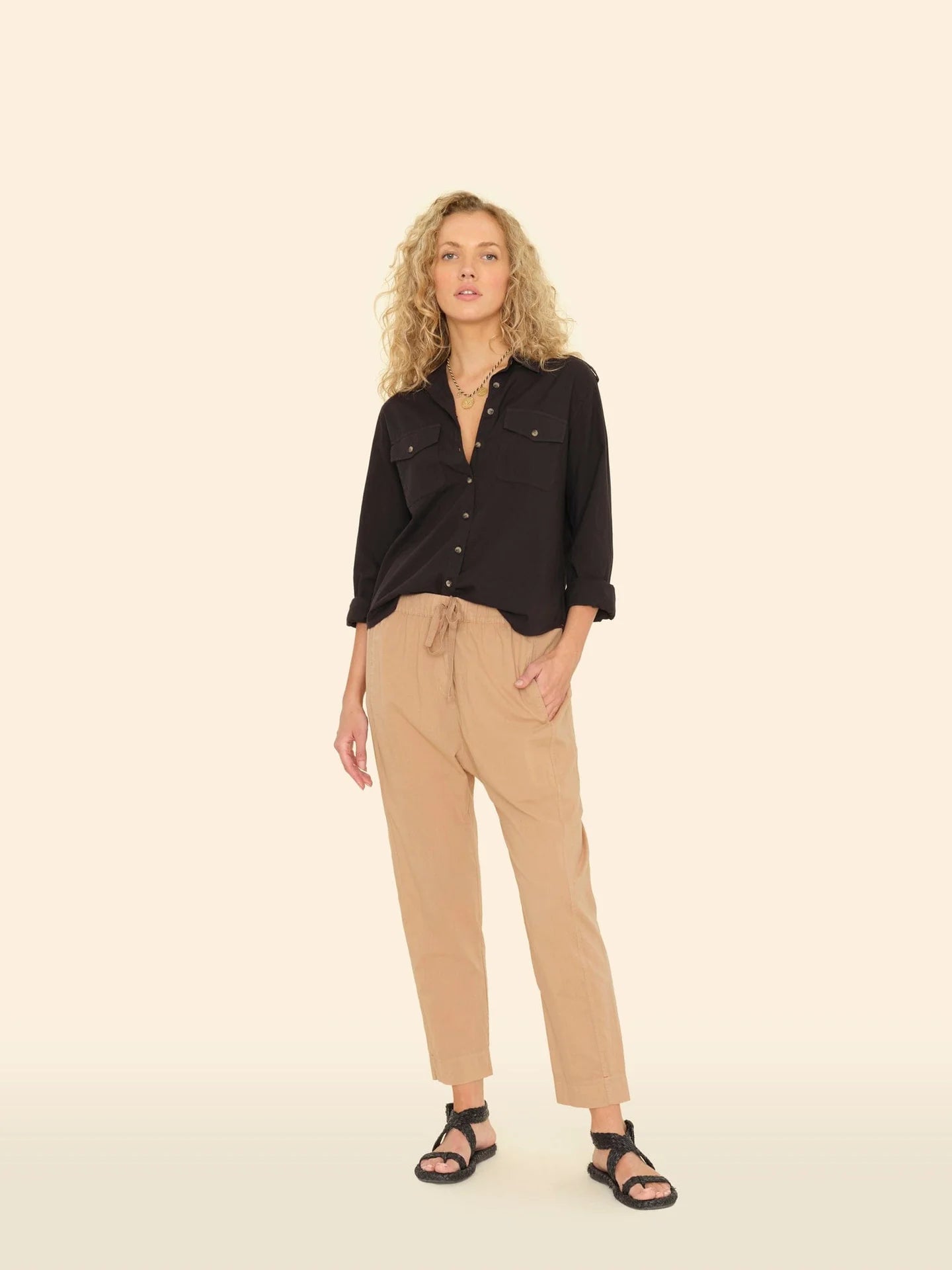 A person with curly blond hair confidently stands against a cream background, wearing a black button-up shirt and Xirena's beige Draper Pants with a drawstring waist. They sport black sandals and look directly at the camera.