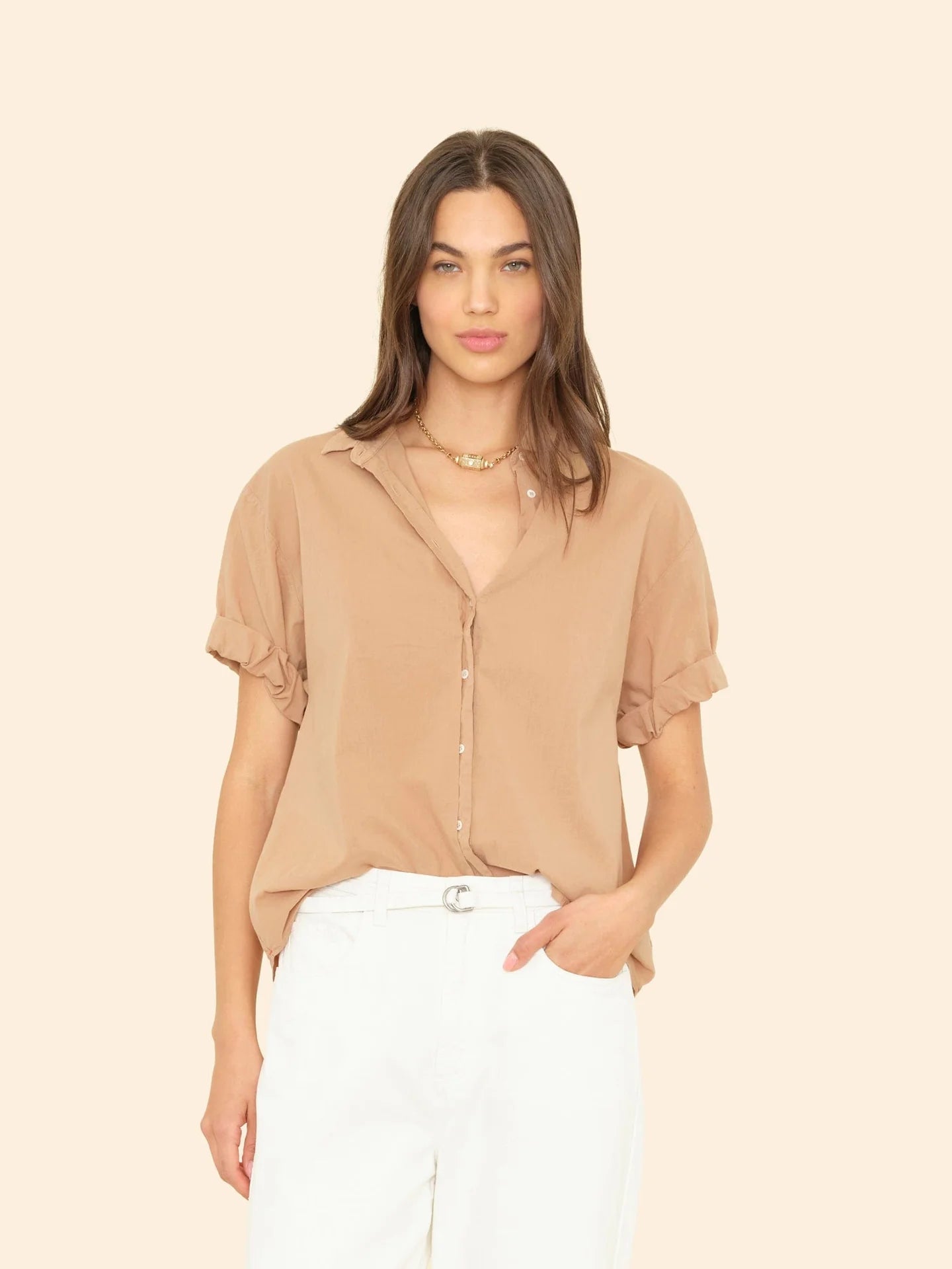 A person with long hair wears a Xirena Channing Shirt made of lightweight cotton poplin, featuring short ruffled sleeves. It's partially tucked into white pants against a soft beige background. They have one hand in their pocket and look forward.