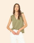 A woman stands against a beige background, looking forward with her hands on her hips, wearing the Xirena Bex Top in olive green cotton poplin and white pants.