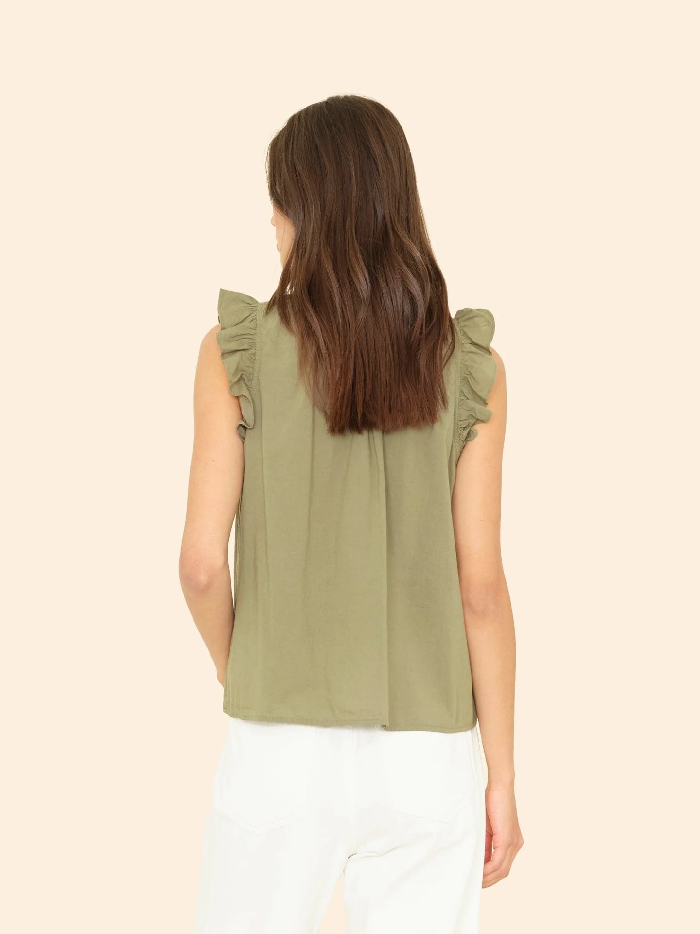 A woman with long brown hair, facing away from the camera, wears the Xirena Bex Top—a sleeveless olive green cotton poplin ruffle popover—with white pants against a plain beige background.