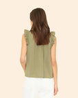 A woman with long brown hair, facing away from the camera, wears the Xirena Bex Top—a sleeveless olive green cotton poplin ruffle popover—with white pants against a plain beige background.