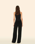 A woman with wavy brown hair, in a black sleeveless top and Xirena’s Delany Pant—wide legged with an elasticized waist—stands facing away from the camera against a plain light beige background.