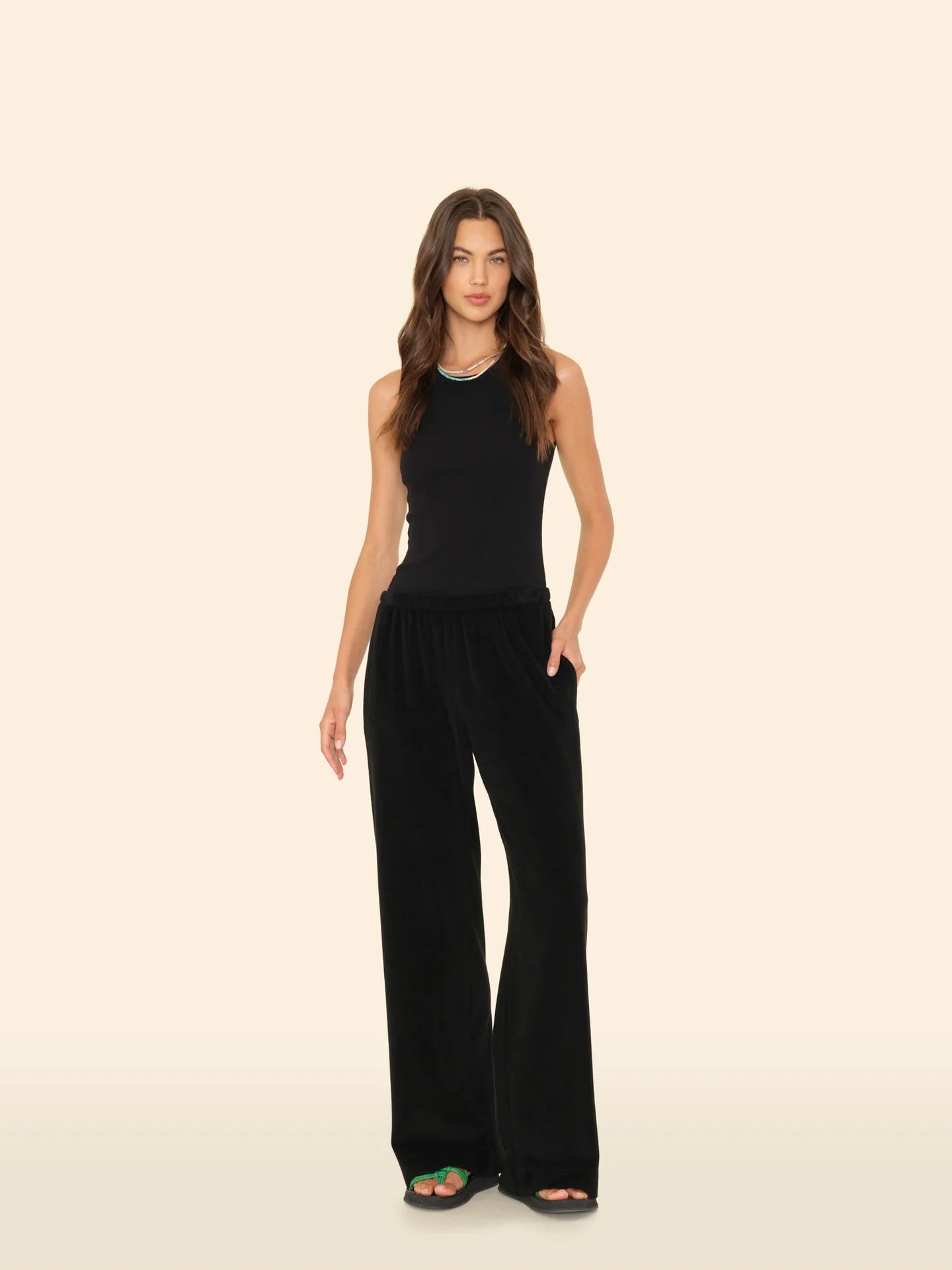 A woman with long, wavy brown hair models the Xirena Delany Pant—loose, wide-leg sweatpants with an elastic waist—paired with a sleeveless black top and black sandals featuring a green accent, standing before a beige backdrop.