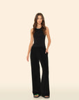 A woman with long, wavy brown hair models the Xirena Delany Pant—loose, wide-leg sweatpants with an elastic waist—paired with a sleeveless black top and black sandals featuring a green accent, standing before a beige backdrop.