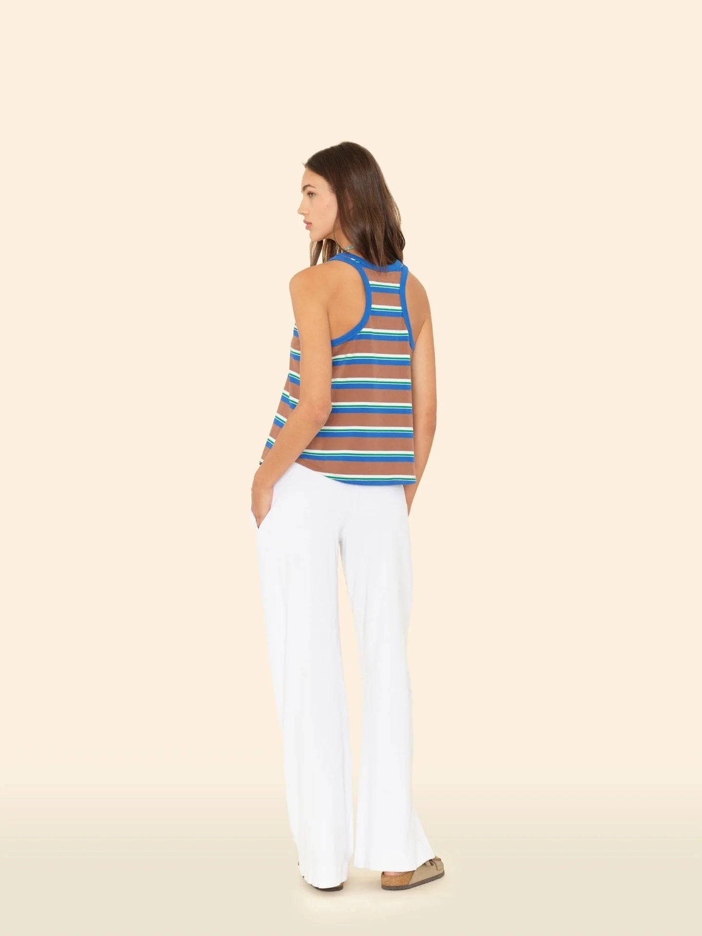 A woman stands with her back turned, wearing the Xirena Delany Pant—cotton blend towel terry wide-leg pants—with a blue and white striped sleeveless top, hands in pockets, against a plain beige background.