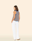 A woman stands with her back turned, wearing the Xirena Delany Pant—cotton blend towel terry wide-leg pants—with a blue and white striped sleeveless top, hands in pockets, against a plain beige background.