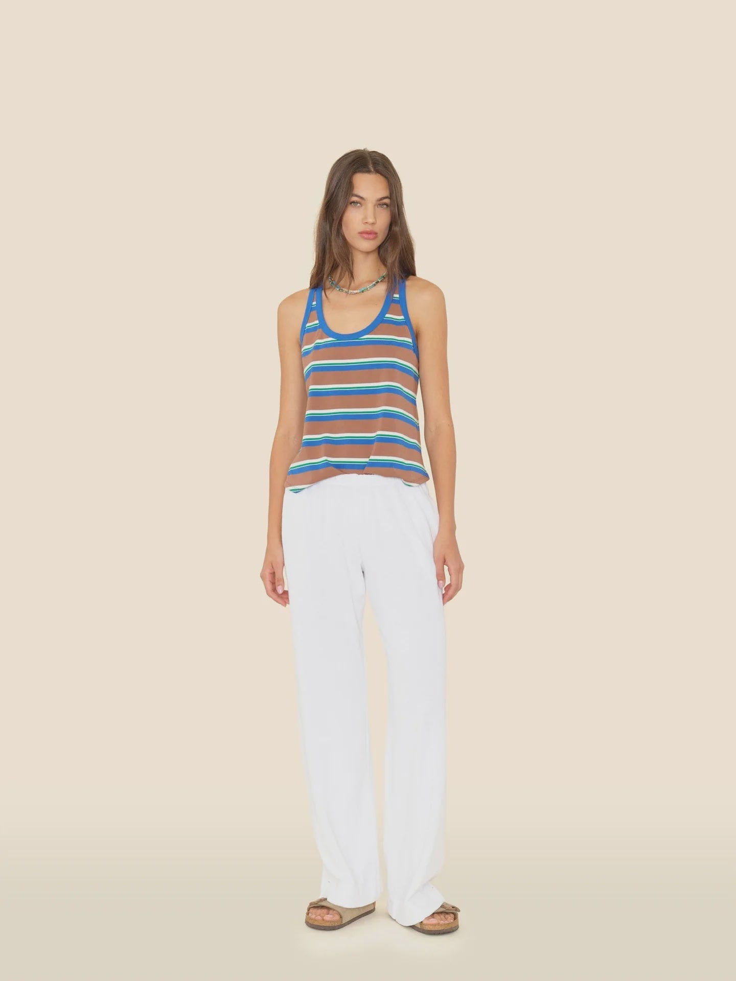 A woman stands facing forward wearing the Xirena Delany Pant—loose, white, wide-leg pants—paired with a blue and brown striped towel terry tank top and sandals against a plain beige background.