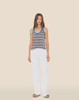 A woman stands facing forward wearing the Xirena Delany Pant—loose, white, wide-leg pants—paired with a blue and brown striped towel terry tank top and sandals against a plain beige background.
