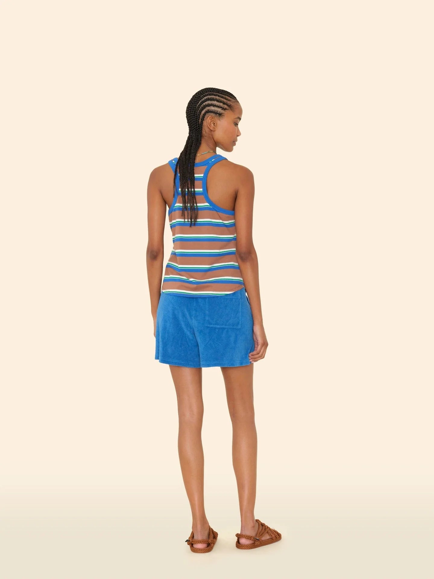 A woman with braided hair stands against a beige background, wearing the Xirena Shayden Short in bright blue and a striped sleeveless top with brown sandals.