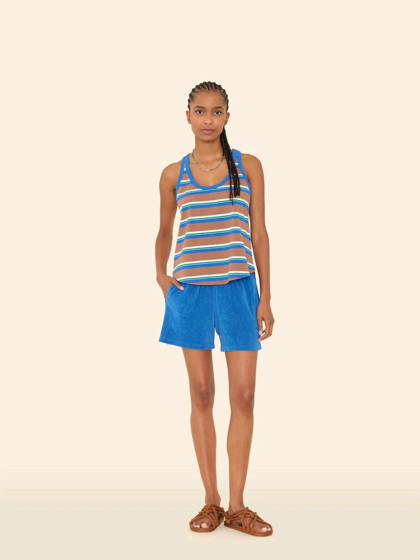 A woman with braided hair stands against a light beige background, wearing the Xirena Shayden Short in bright blue with an elasticized waist, a cotton blend striped tank top, and brown sandals. She looks at the camera with one hand in her pocket.