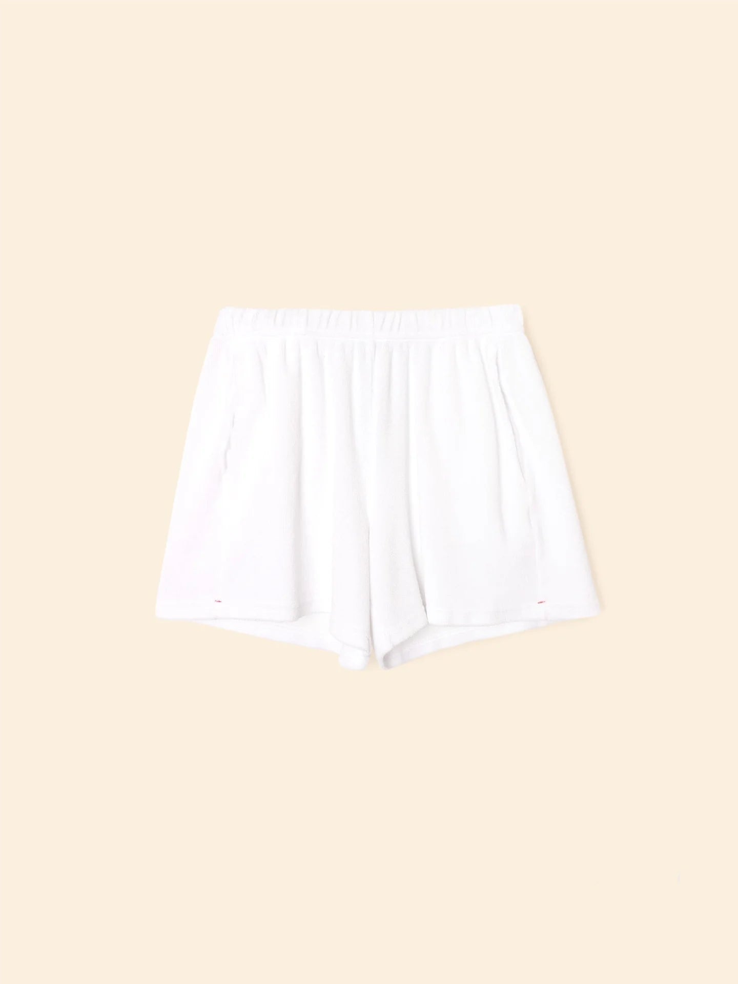 The Shayden Short by Xirena is a white terry sweat short with an elastic waist and side pockets, made from a soft cotton blend and shown flat on a beige background.