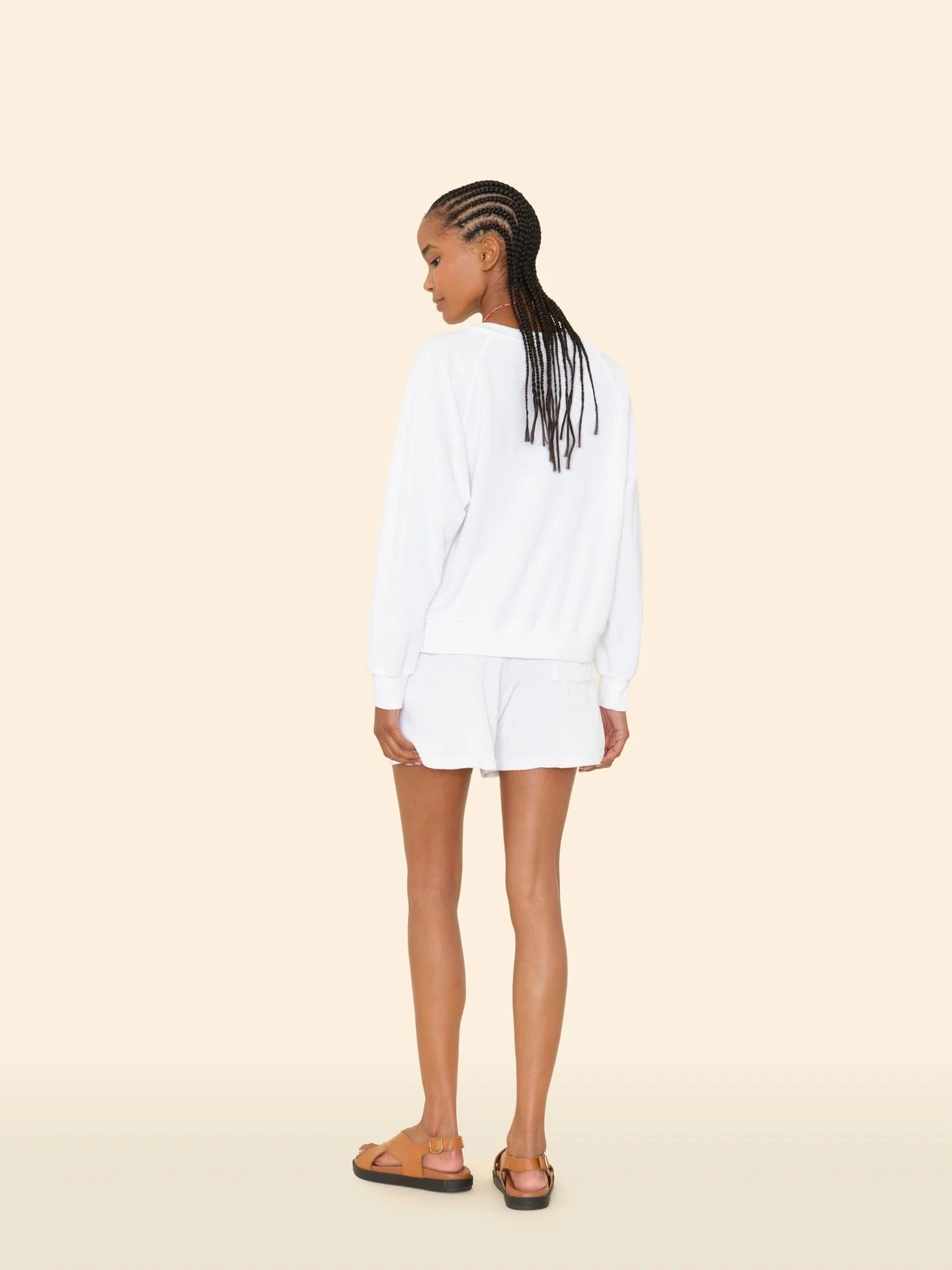 A woman with braided hair stands facing away, wearing a white cotton blend long-sleeve top and Xirena’s Shayden Short, featuring an elasticized waist, paired with tan sandals against a plain beige background.