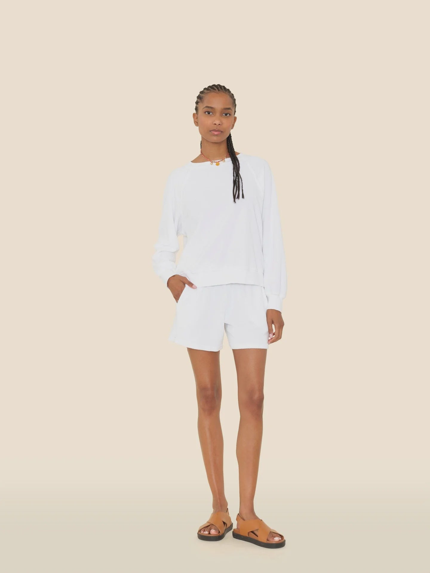 A woman with braided hair poses against a beige background in a white long-sleeve shirt, Xirena Shayden Shorts with an elastic waist, brown sandals, and a brown necklace. One hand is in her pocket.