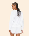 A woman with long, braided hair stands facing away, wearing the Xirena Hopper Sweatshirt and matching white shorts in a cotton-blend terry fabric against a light beige background.