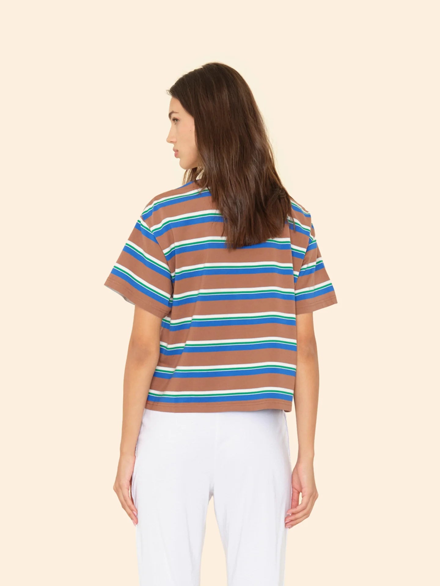 A woman with long brown hair, seen from behind, wears the Xirena Paulina Tee—a striped cotton jersey in blue, white, and yellow with a contrast ringer neck—paired with white pants against a plain beige background.