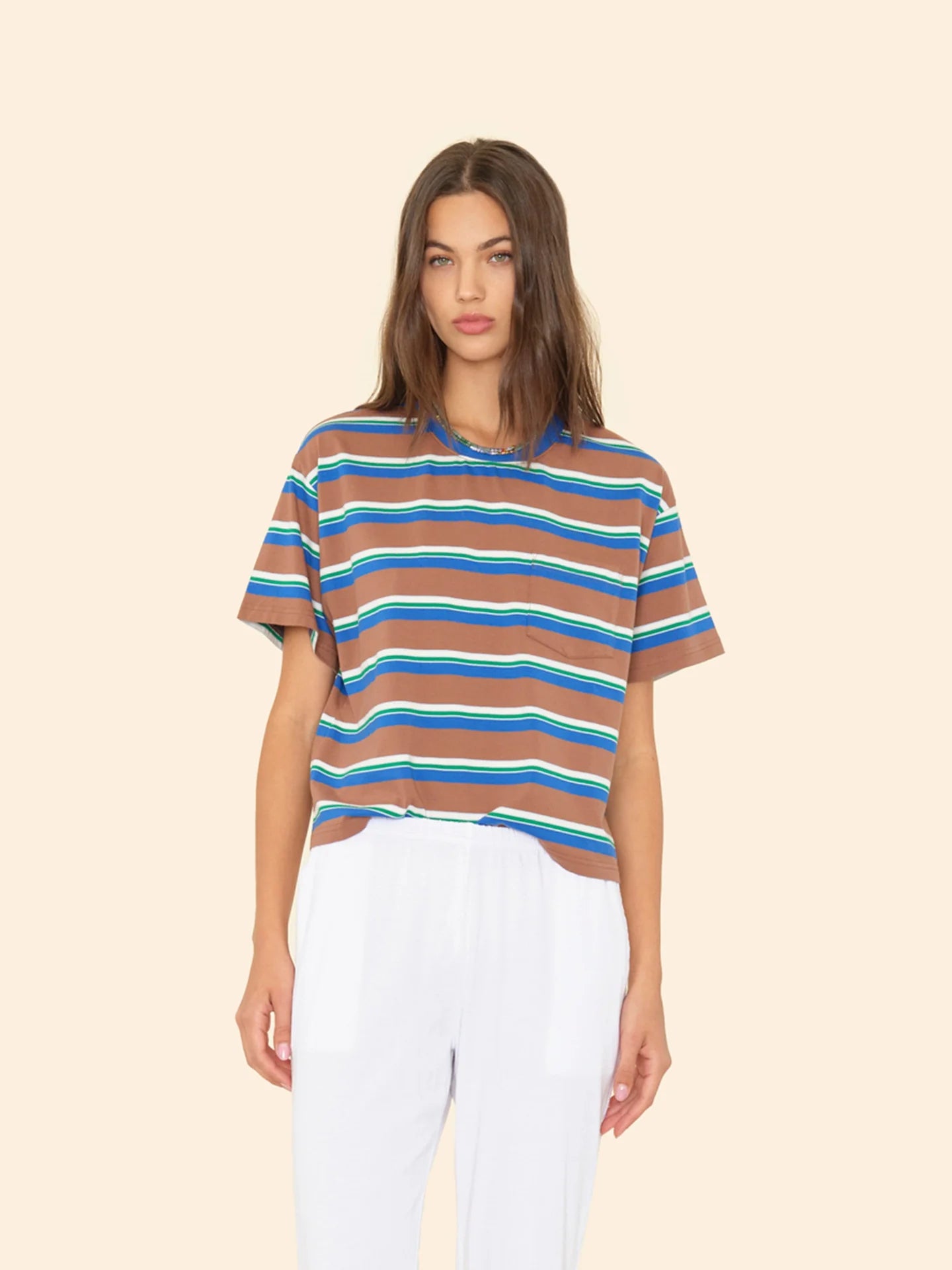 A woman with long brown hair wears the Xirena Paulina Tee in blue and white striped cotton jersey with a contrast ringer neck, paired with white pants, standing against a plain beige background.
