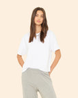 A woman with long brown hair wears the Xirena Palmer Tee, a loose cotton jersey wide-sleeve top, and light gray sweatpants, standing against a plain beige background with a relaxed expression.