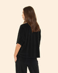 A woman with long brown hair is shown from the side and back, wearing a loose-fitting Xirena Roe Top and black pants, her right hand in her pocket, standing against a plain beige background.