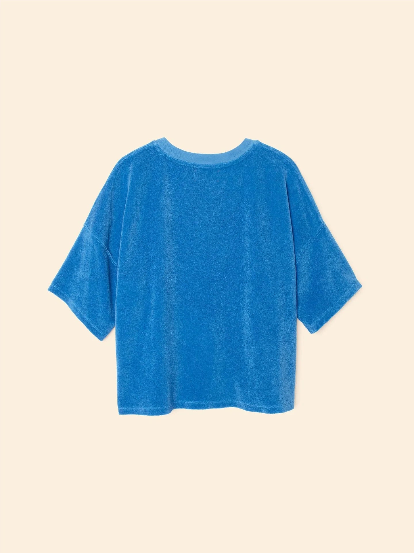 A loose-fitting, short-sleeved blue Roe Top by Xirena is laid flat on a beige background and shown from the back.