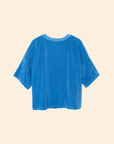 A loose-fitting, short-sleeved blue Roe Top by Xirena is laid flat on a beige background and shown from the back.