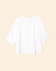 The Roe Top by Xirena, an oversized plain white short-sleeve T-shirt in a soft cotton blend, is shown from the back on a light beige background.