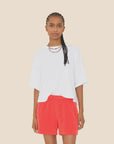 A person with braided hair wears the Roe Top by Xirena, a loose white cotton blend T-shirt, and bright red shorts, standing against a plain beige background.