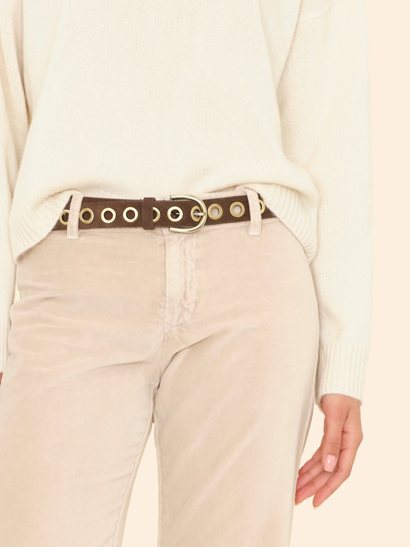 A person wears the Xirena Francoise Belt—a brown leather belt with large gold grommets and an antique brass buckle—over beige pants and a cream sweater. Only the lower torso and one hand are visible.