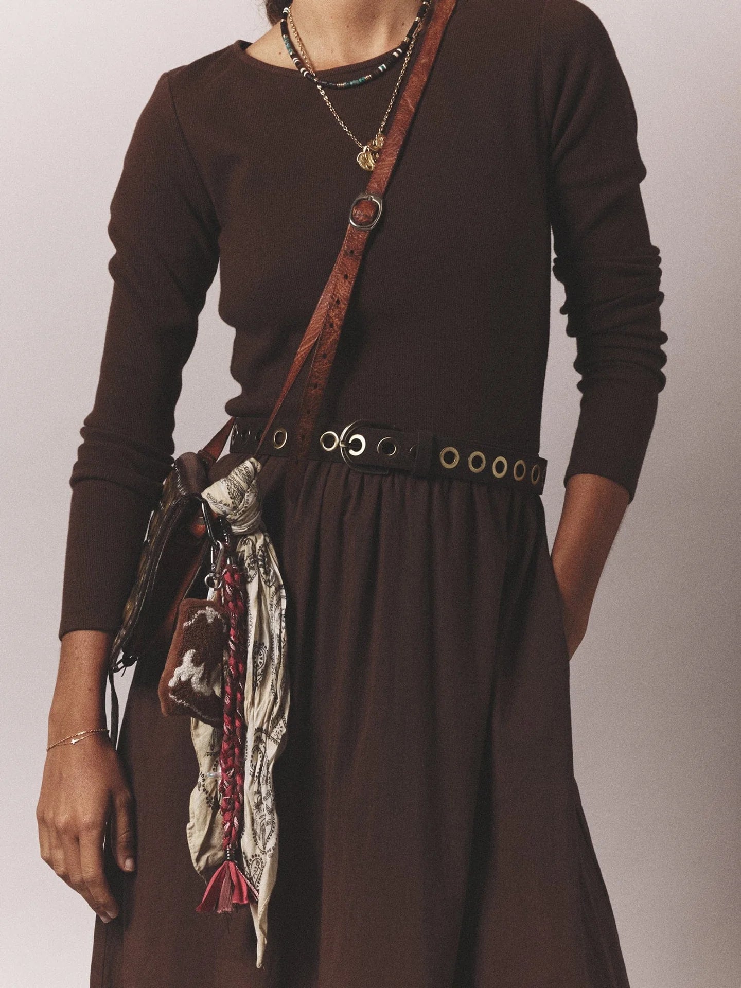 A person wearing a long-sleeve brown dress with the Xirena Francoise Belt—an adjustable black leather belt with an antique brass buckle—layered necklaces, a brown crossbody bag, and a patterned tassel scarf attached to the bag.
