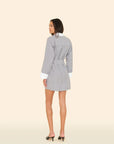 A woman stands facing away, looking over her shoulder. She wears the Xirena Louie Dress—a belted striped cotton style with white cuffs and collar—paired with black heeled sandals against a solid light beige background.