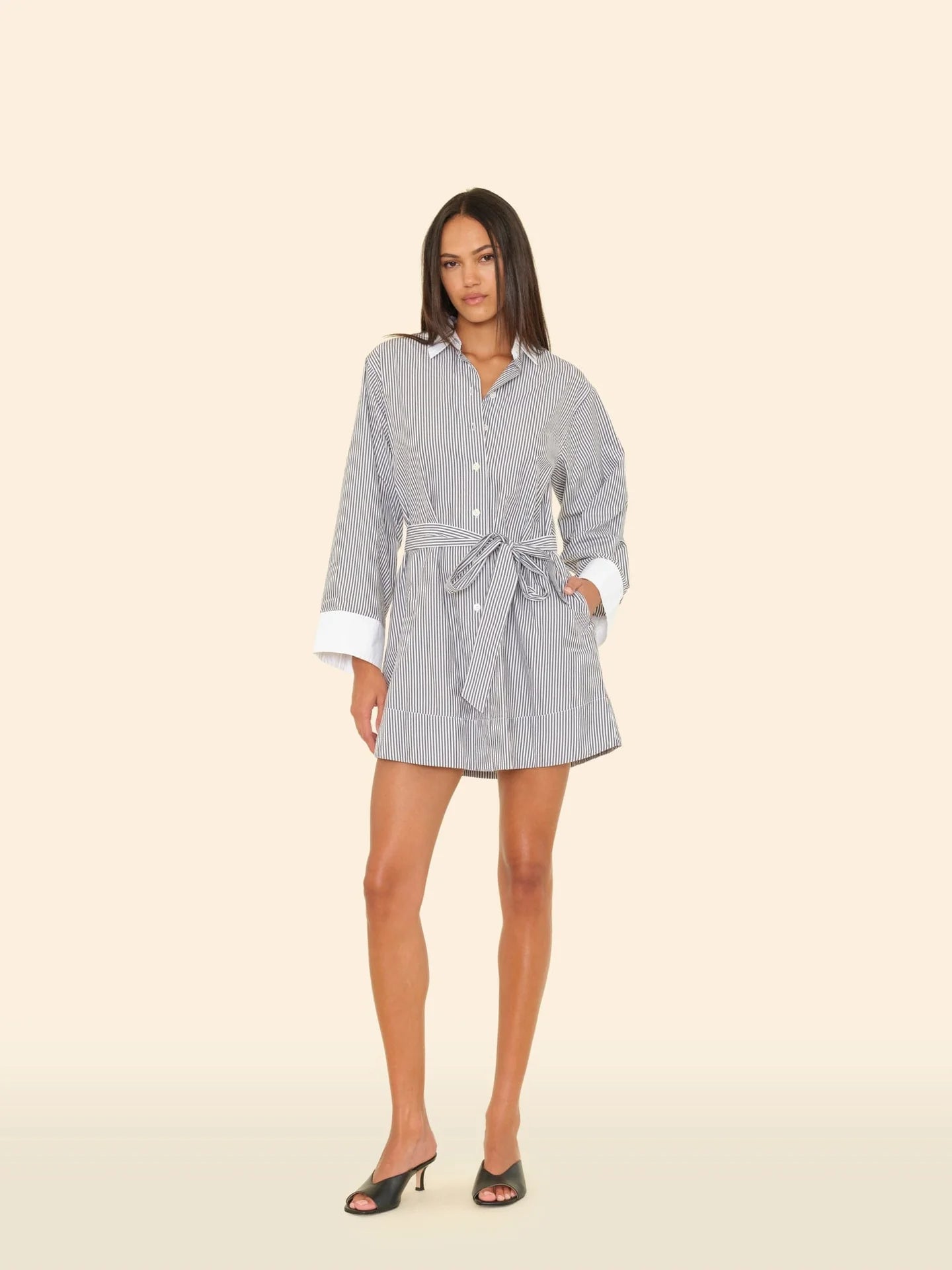 A woman with long, straight dark hair wears the Xirena Louie Dress—a multicolor stripe cotton mini shirtdress with white cuffs and a removable self-tie belt—paired with black open-toe heeled mules against a plain beige background.