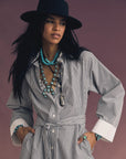 A woman poses confidently against a muted mauve background, wearing the Xirena Louie Dress—a black-and-white striped mini shirtdress—paired with a black wide-brimmed hat and multiple turquoise jewelry pieces.